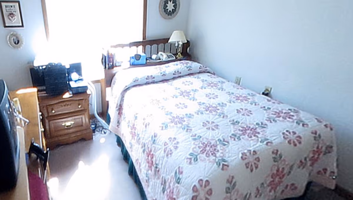 Small bedroom with a floral quilted bedspread, wooden headboard, nightstand with lamp and a TV on a dresser.