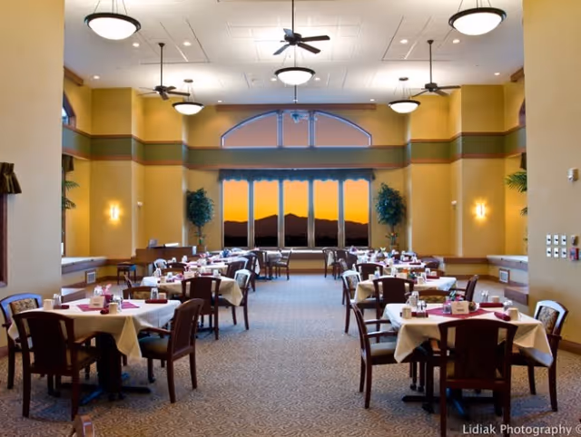A spacious dining room with multiple tables covered in white tablecloths and set with napkins and condiments. The room has high ceilings with ceiling fans and round light fixtures. Large windows at the far end show a scenic view of mountains during sunset. The walls are painted in warm tones and there are potted plants near the windows.