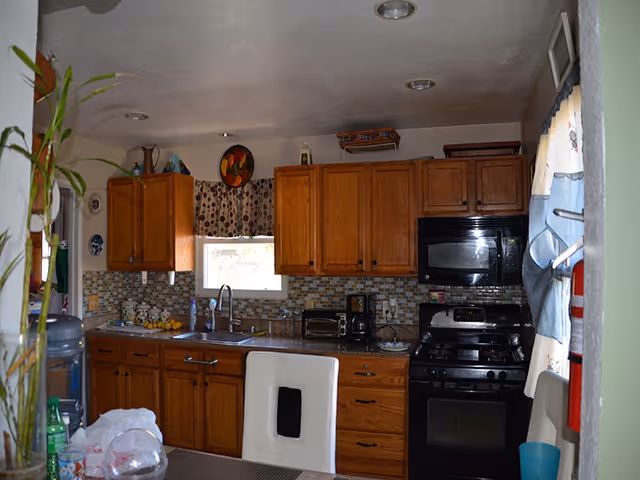 Interior view of a kitchen with wooden cabinets, a tiled backsplash, a window with patterned curtains above the sink, a black microwave and stove, a toaster, coffee maker, and various kitchen items on the countertop. A white chair is partially visible in the foreground, and a water dispenser and some plants are on the left side.