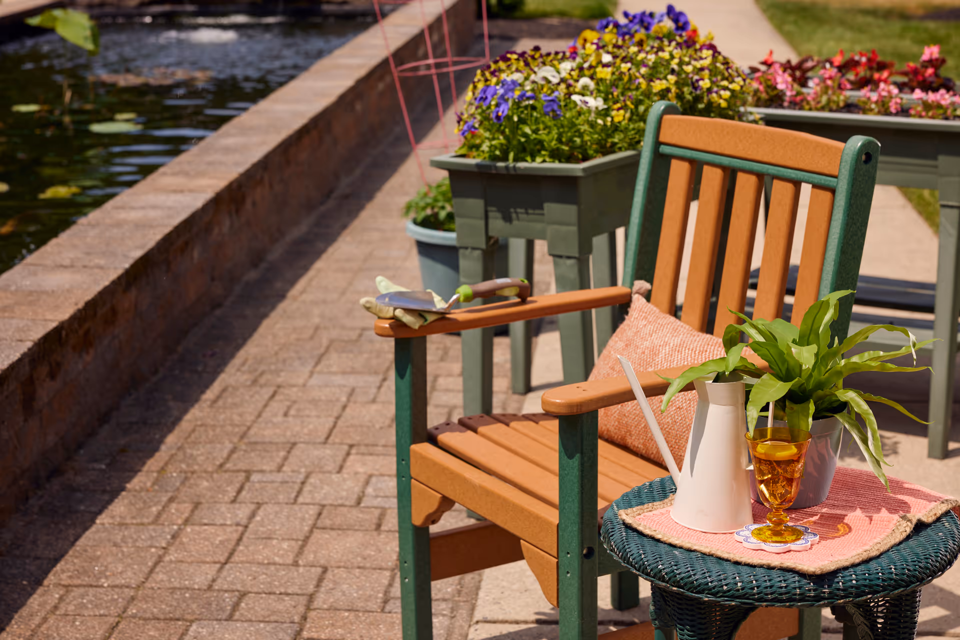A wooden chair with green metal frame and a pink cushion is placed on a paved pathway next to a pond. On a small round wicker table beside the chair, there is a white watering can, a green potted plant, and a glass with an amber-colored drink. In the background, there are flower pots with colorful flowers along the pathway.