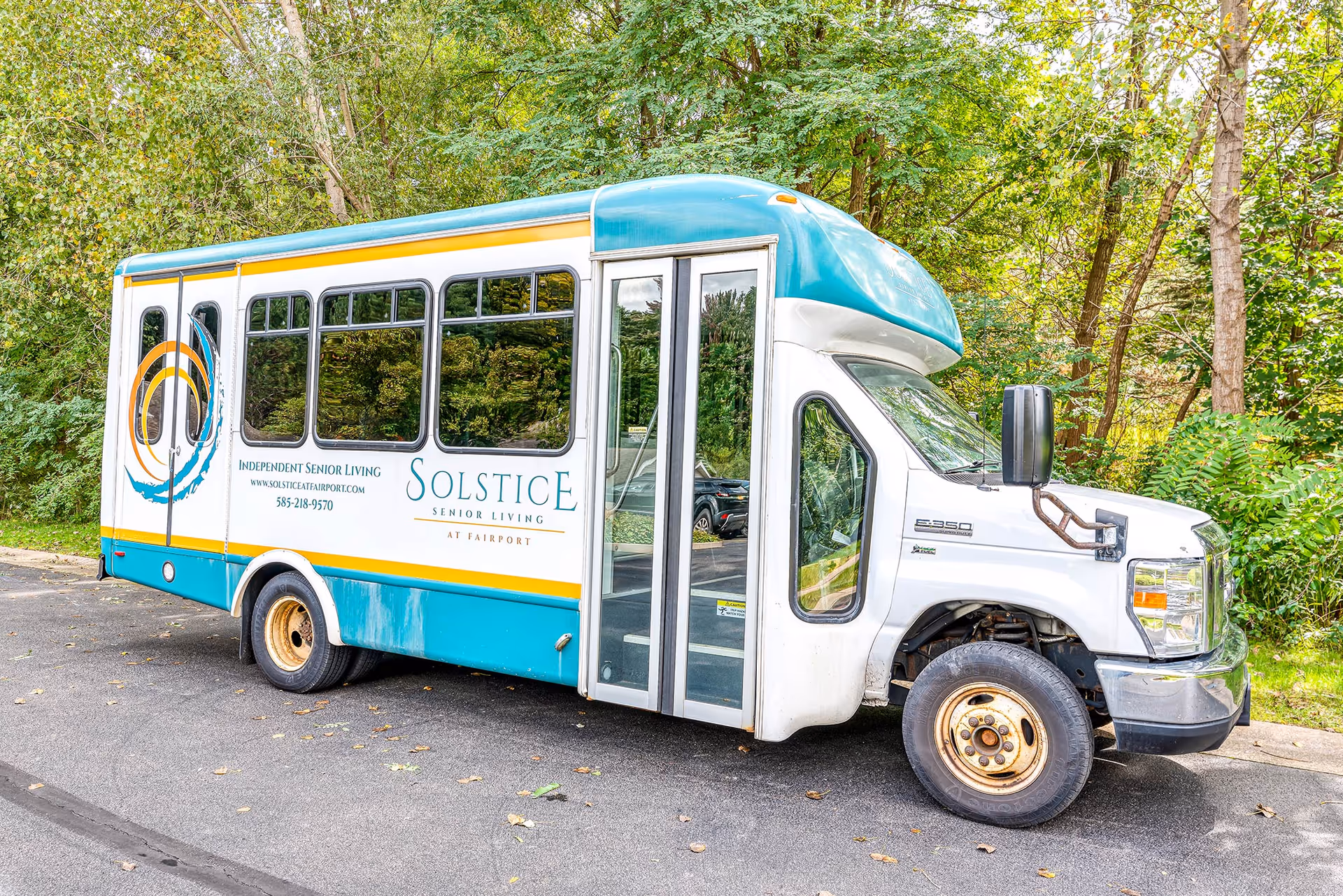 A teal-and-white Solstice Senior Living shuttle bus parked on a tree-lined road.