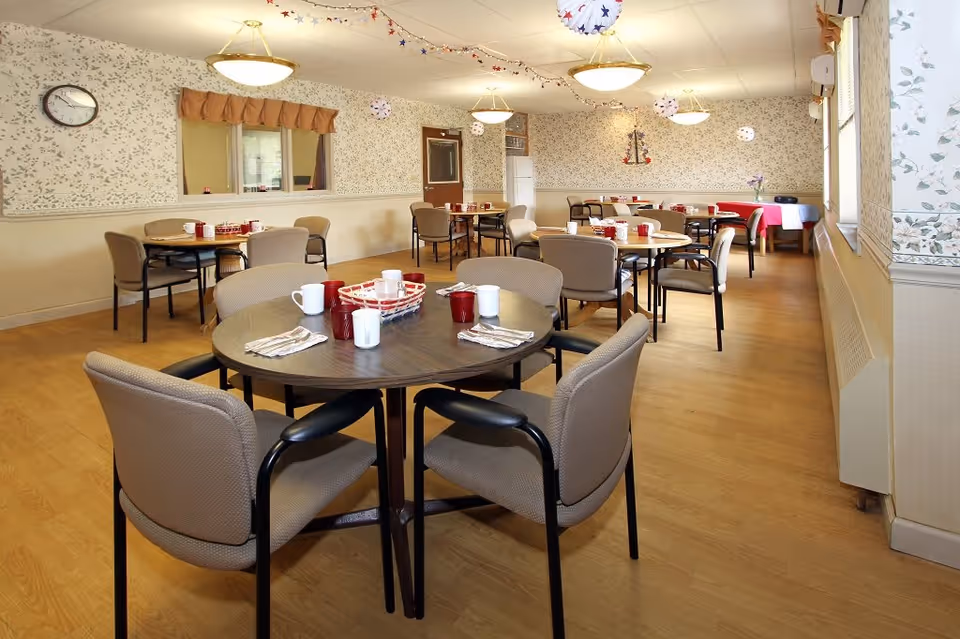 A dining room with several round tables and beige cushioned chairs arranged around them. Each table is set with red and white cups, napkins, and utensils. The room has light wood flooring, floral wallpaper, and ceiling lights. There are patriotic decorations hanging from the ceiling and a clock on the wall.