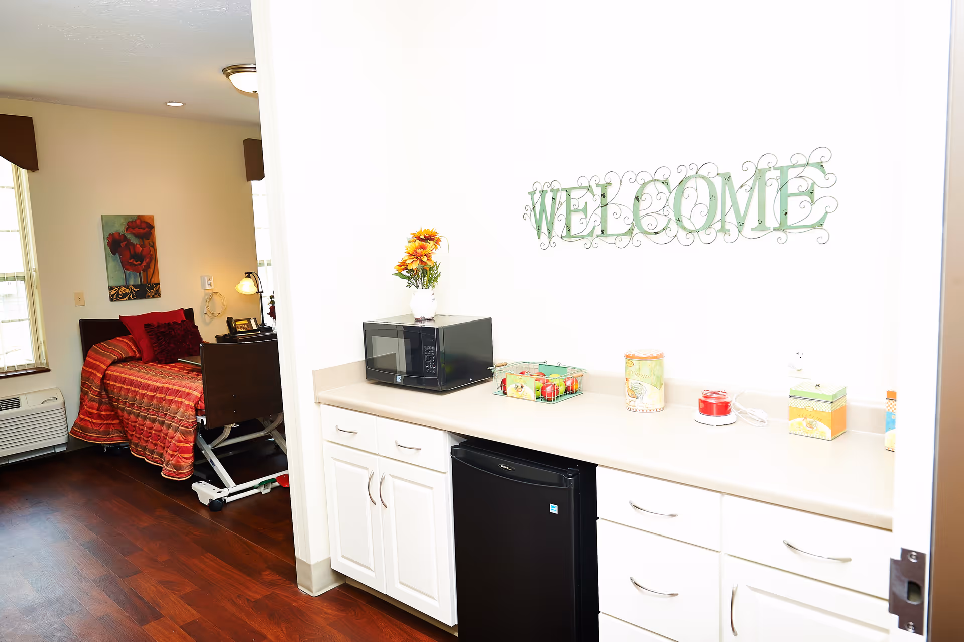Interior view of a senior living facility room showing a kitchenette area with white cabinets, a small black refrigerator, a microwave, and decorative items on the countertop. A green decorative sign on the wall reads 'WELCOME'. In the background, there is a bedroom with a bed covered in a red and orange patterned quilt, a bedside table with a lamp, and a window with brown valances.