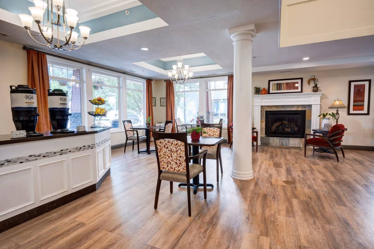 A bright and inviting common area in South Hill Village featuring wooden flooring, several tables with floral upholstered chairs, large windows with orange curtains letting in natural light, a white column, a fireplace with decorative mantle, and two beverage dispensers labeled '1850' on a counter.