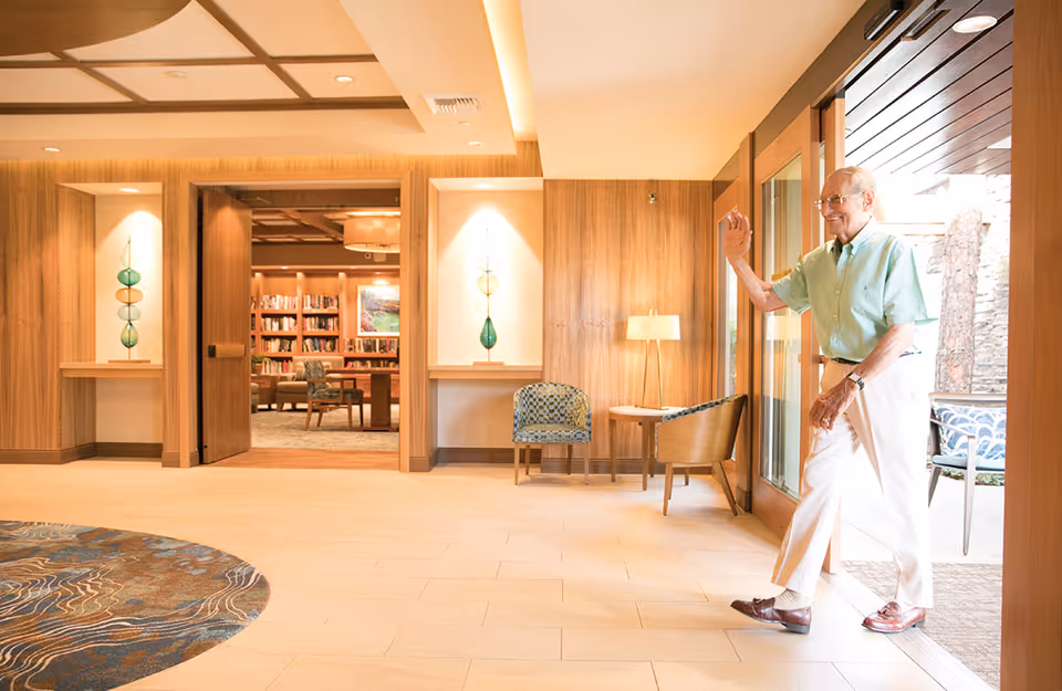 An elderly man walks into a warm, wood-paneled lobby with seating and a view into a library.