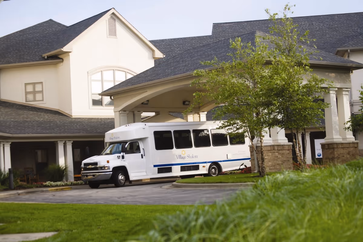 White Village Shalom shuttle parked under the covered entrance of a senior living building with trees and a lawn in front.
