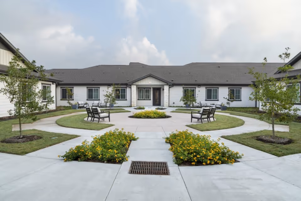 Outdoor courtyard area of a senior living facility with a circular concrete pathway, green grass, small trees, yellow flowering plants, and several benches arranged around the circle. The building surrounding the courtyard has white siding and multiple windows under a dark shingled roof.