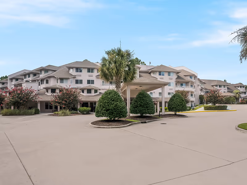 Exterior view of a multi-story senior living facility with light-colored walls and multiple windows. The building is surrounded by neatly trimmed bushes, palm trees, and a wide driveway under a clear blue sky.