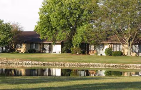 Single-story retirement community buildings with green lawns, trees, and a reflecting pond in front.