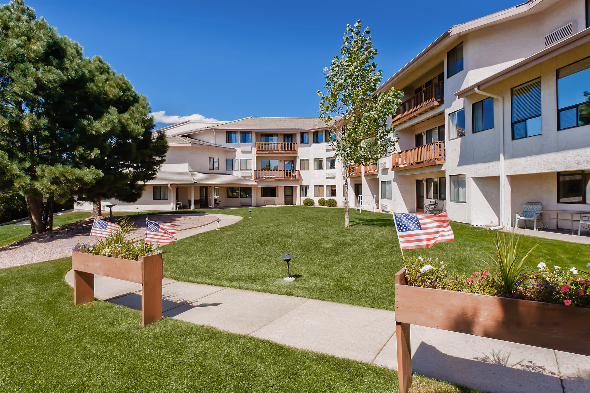 Exterior courtyard of a multi-story senior living building with a manicured lawn, planters displaying American flags, and balconies.