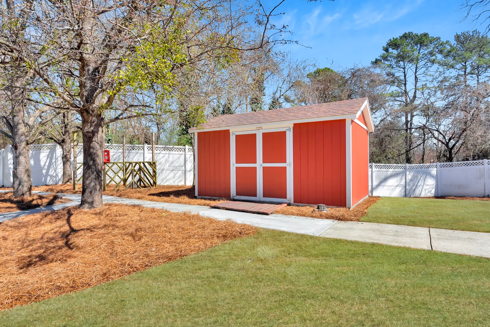 A red wooden shed with white trim situated in a fenced yard with green grass, trees, and a concrete pathway leading to the shed. The yard is surrounded by a white fence and there is a fire extinguisher mounted on a post near the trees.