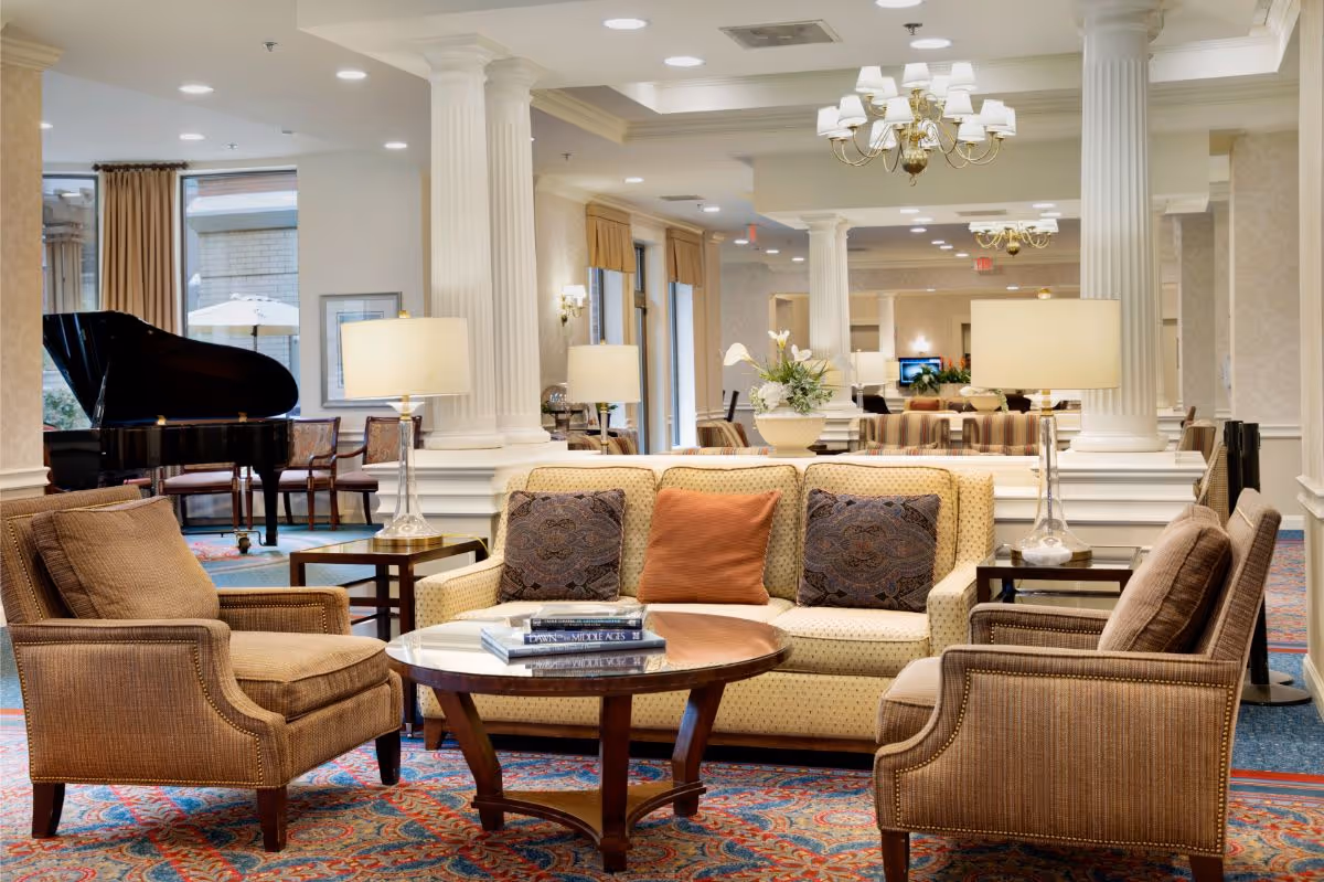 Elegant senior living lounge with sofas, armchairs, a coffee table, table lamps, and a grand piano in the background.