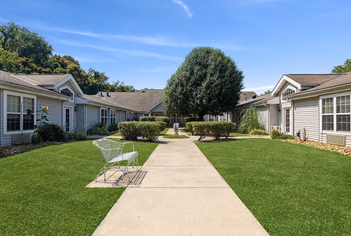 Outdoor courtyard area at The Addison of Uniontown with a concrete walkway, green grass, trimmed bushes, a large tree in the center, and a white metal bench on the left side. The courtyard is surrounded by single-story buildings with windows and doors under a clear blue sky.