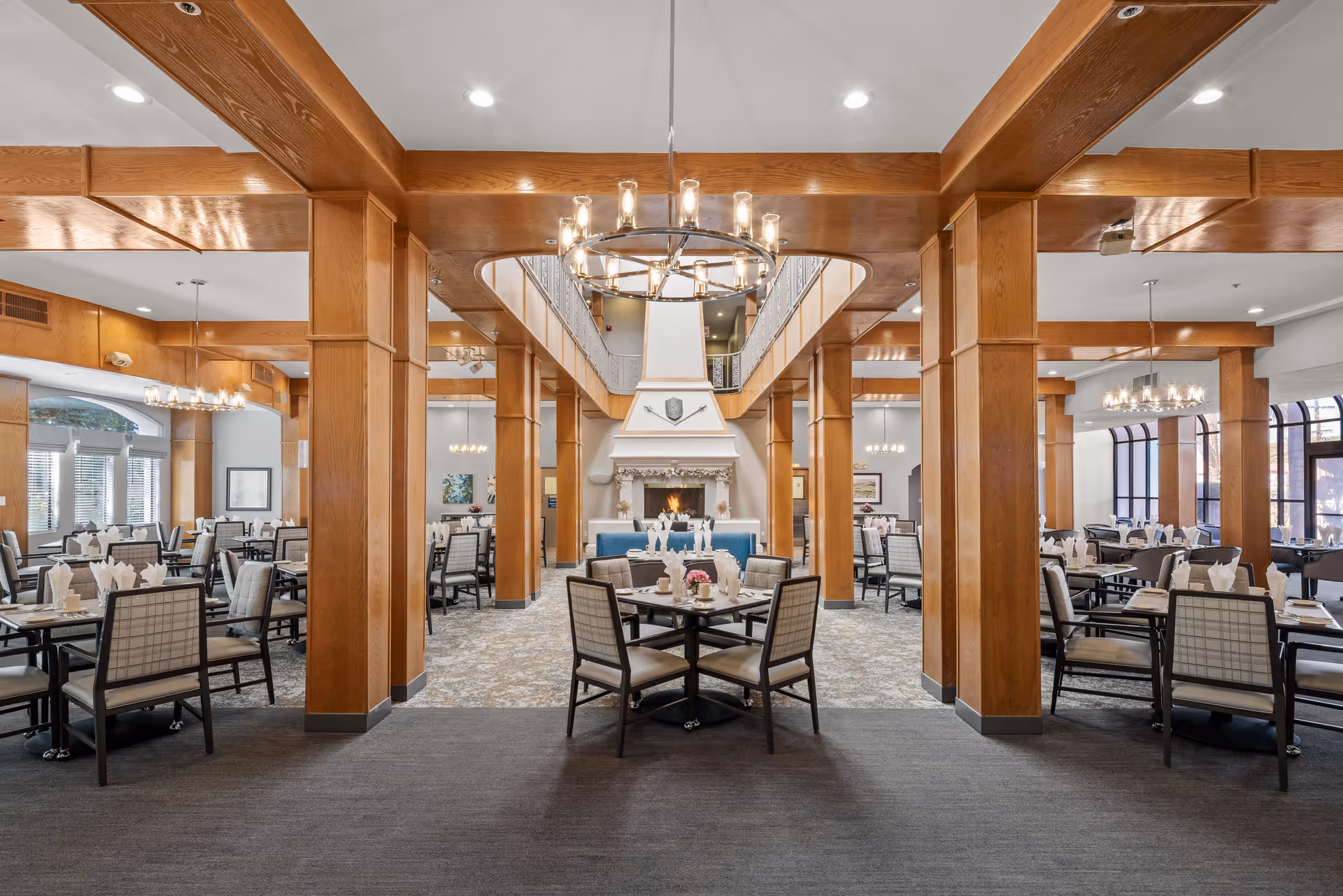 Spacious dining room in a senior living community with multiple tables and chairs arranged neatly. The room features wooden beams and columns, a large chandelier, and a central fireplace with a decorative mantle. Large windows allow natural light to fill the space.