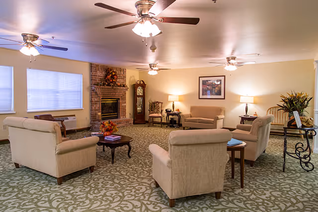A cozy living room area with beige upholstered armchairs and sofas arranged around a wooden coffee table. The room features a patterned carpet, a brick fireplace decorated with autumn-themed foliage, a grandfather clock, and several table lamps providing warm lighting. Ceiling fans with lights are mounted on the ceiling, and large windows with blinds allow natural light to enter.
