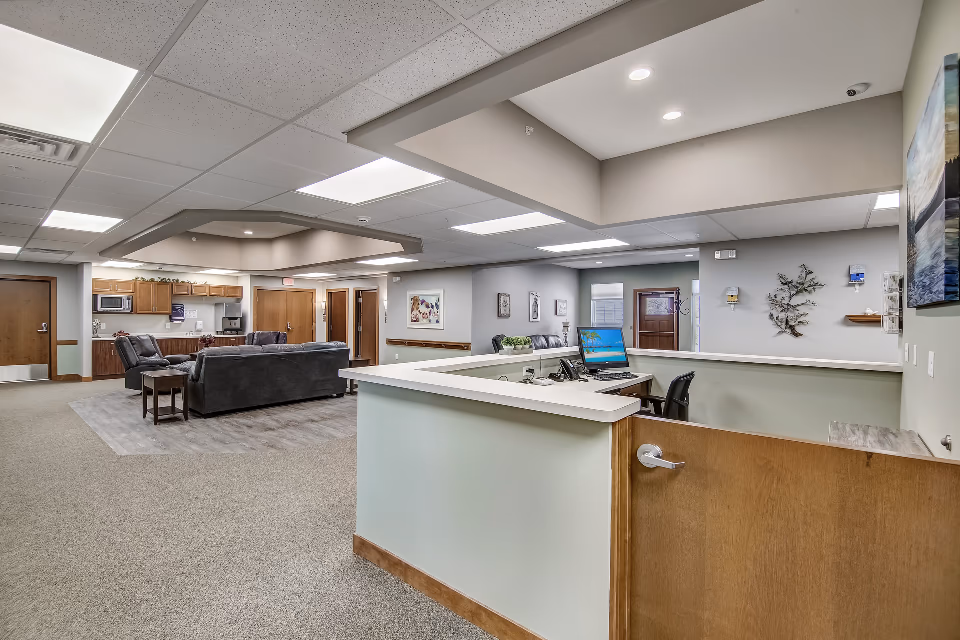 Reception desk and lounge area with sofas, a kitchenette, and workstations inside a senior living facility.