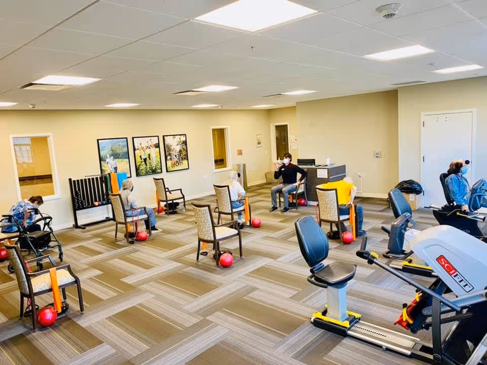 A senior living facility exercise room with several elderly individuals seated on chairs spaced apart, engaging in light exercise activities with red balls and resistance bands. The room has exercise equipment including stationary bikes, beige walls, framed pictures on the wall, and a patterned carpeted floor.