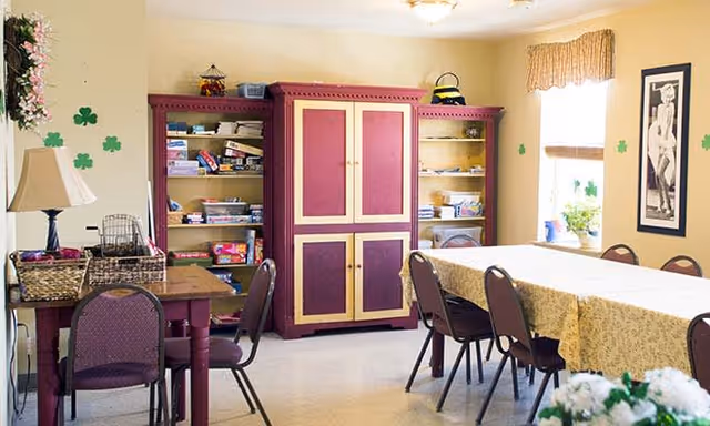 A well-lit room with a long table covered with a yellow patterned tablecloth surrounded by chairs. There is a red and yellow cabinet with shelves filled with board games and other items. A smaller table with chairs and a lamp is on the left side. The walls are decorated with green shamrock cutouts and a framed black and white photograph. A window with a valance lets in natural light.