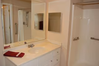 Bathroom with a white vanity featuring a sink and faucet, a large mirror above the sink, a red towel folded on the countertop, and a walk-in shower with grab bars.