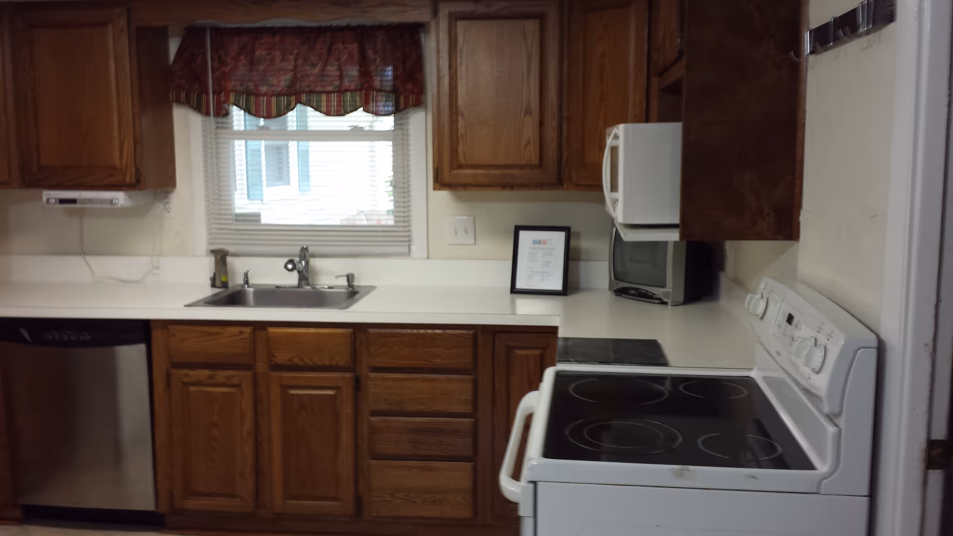 A kitchen with wooden cabinets, a white electric stove, a microwave, a stainless steel dishwasher, a sink under a window with blinds and a red patterned valance.