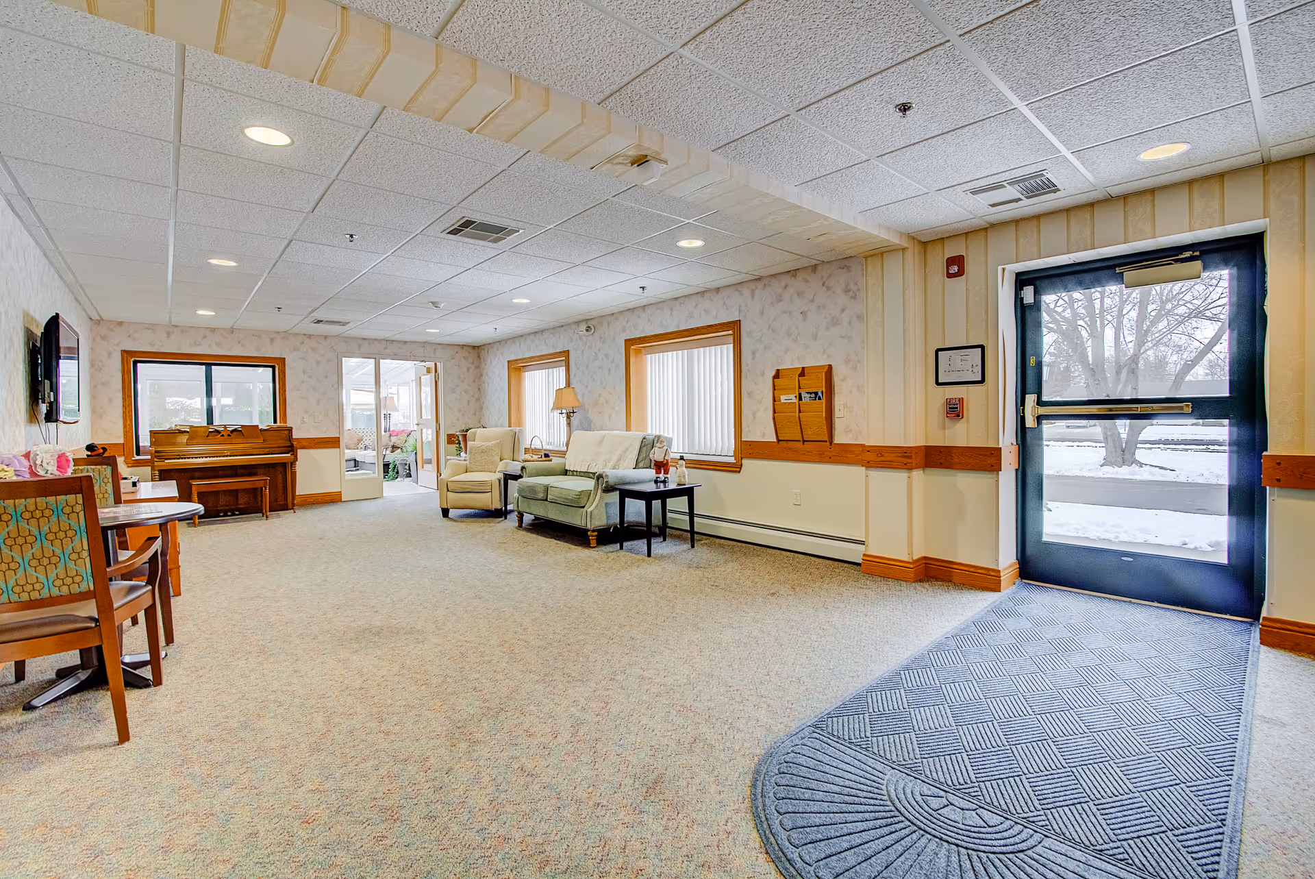 Spacious senior living common room with chairs, a sofa, a piano and a glass exit door showing a snowy yard.