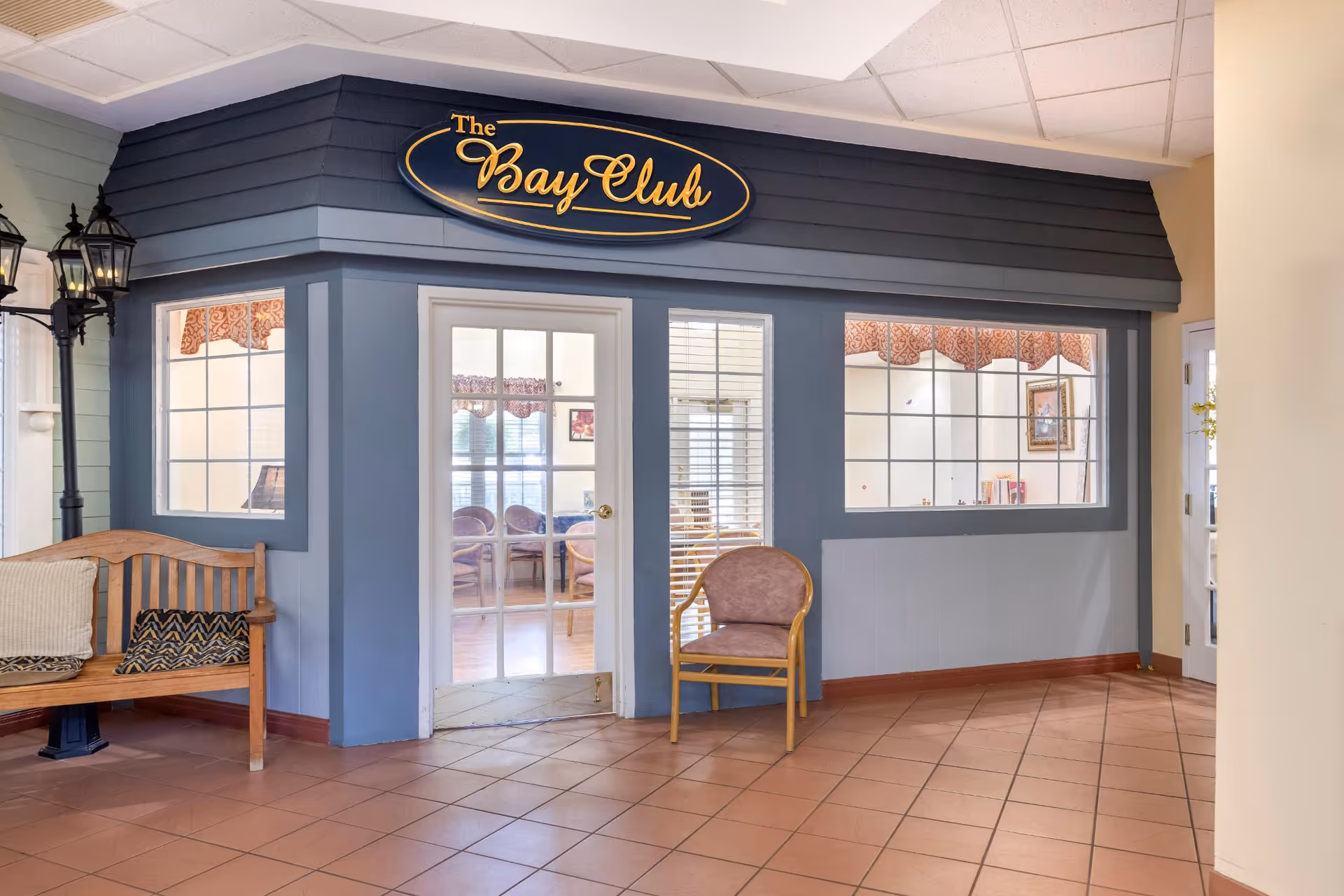 Interior view of a common area named The Bay Club in a senior living facility. The area features a blue-gray wall with large windows and a glass door, a wooden bench with cushions on the left, a single chair on the right, and a tiled floor. There are decorative street lamp-style lights on the left side.
