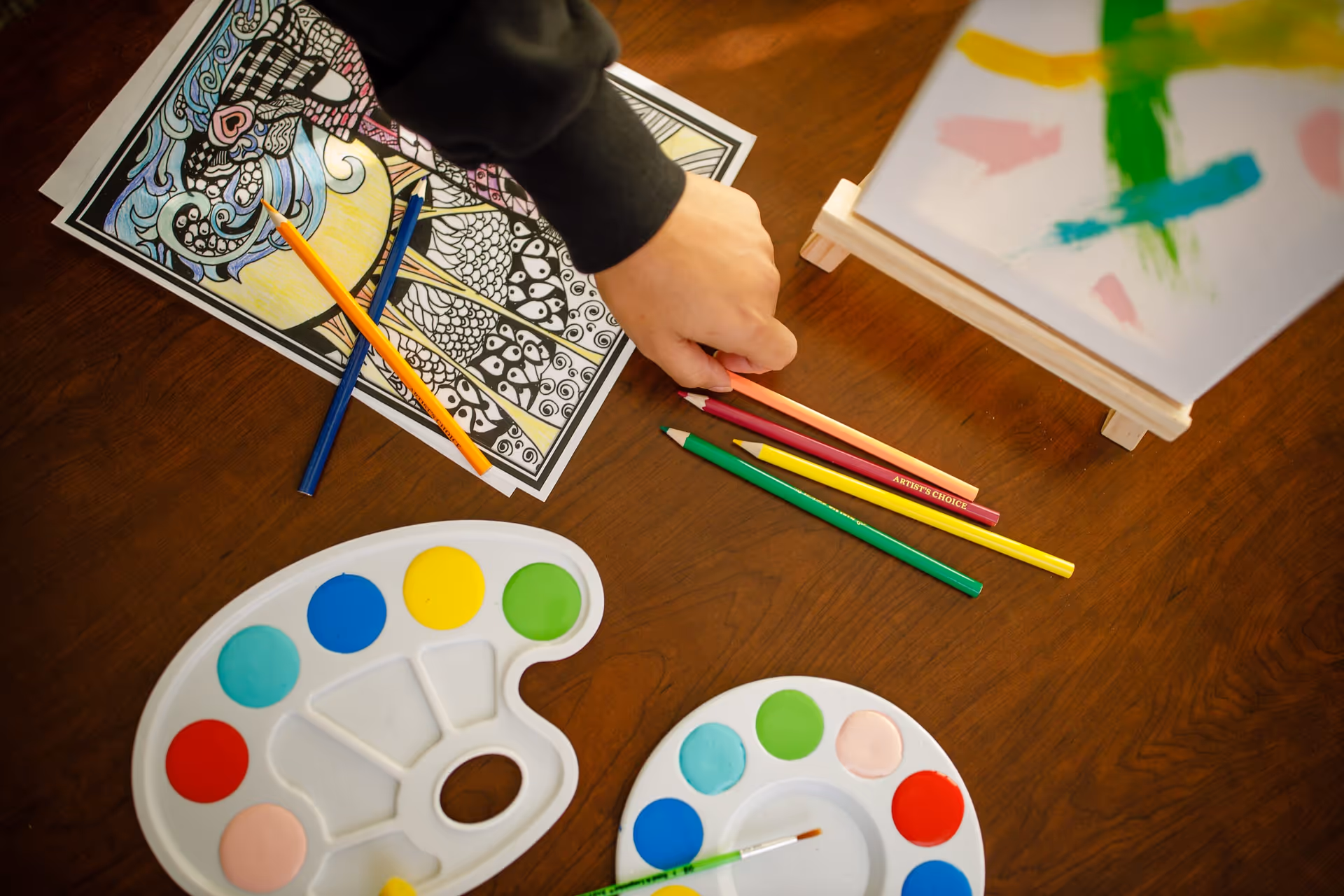A hand reaching for colored pencils on a wooden table with two paint palettes, a paintbrush, a partially colored intricate drawing, and a small easel holding a canvas with abstract paint strokes.