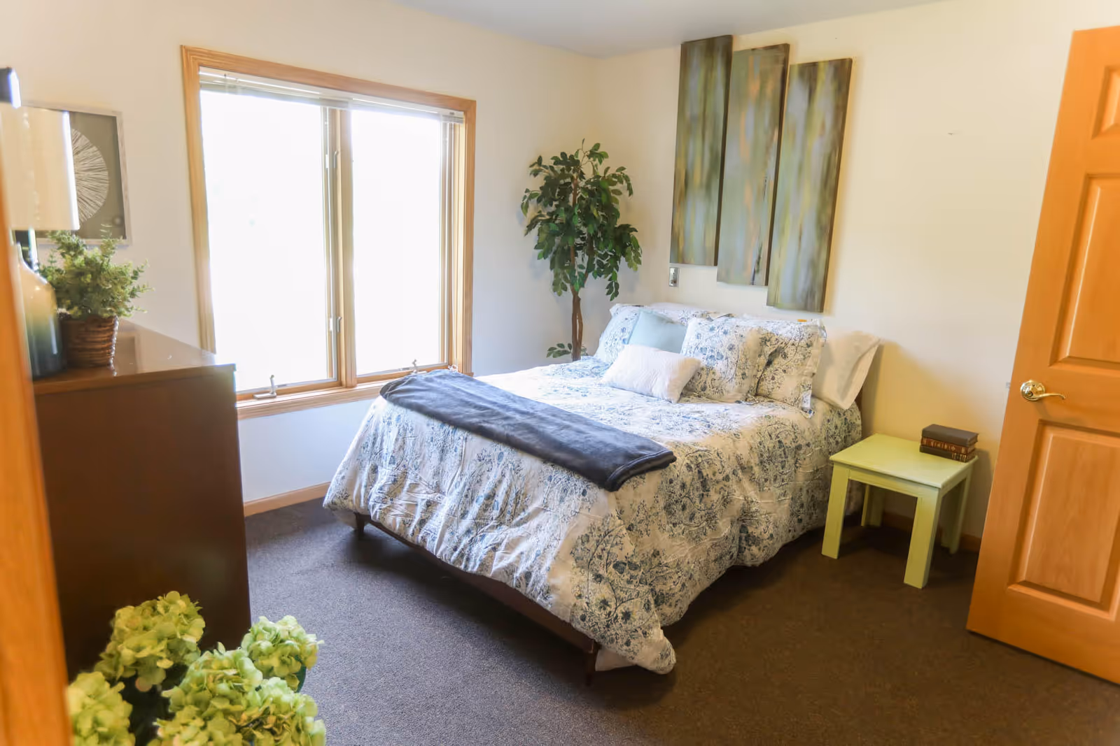 A cozy sunlit bedroom with a double bed in patterned bedding, a window, dresser, potted plant, and a small nightstand.
