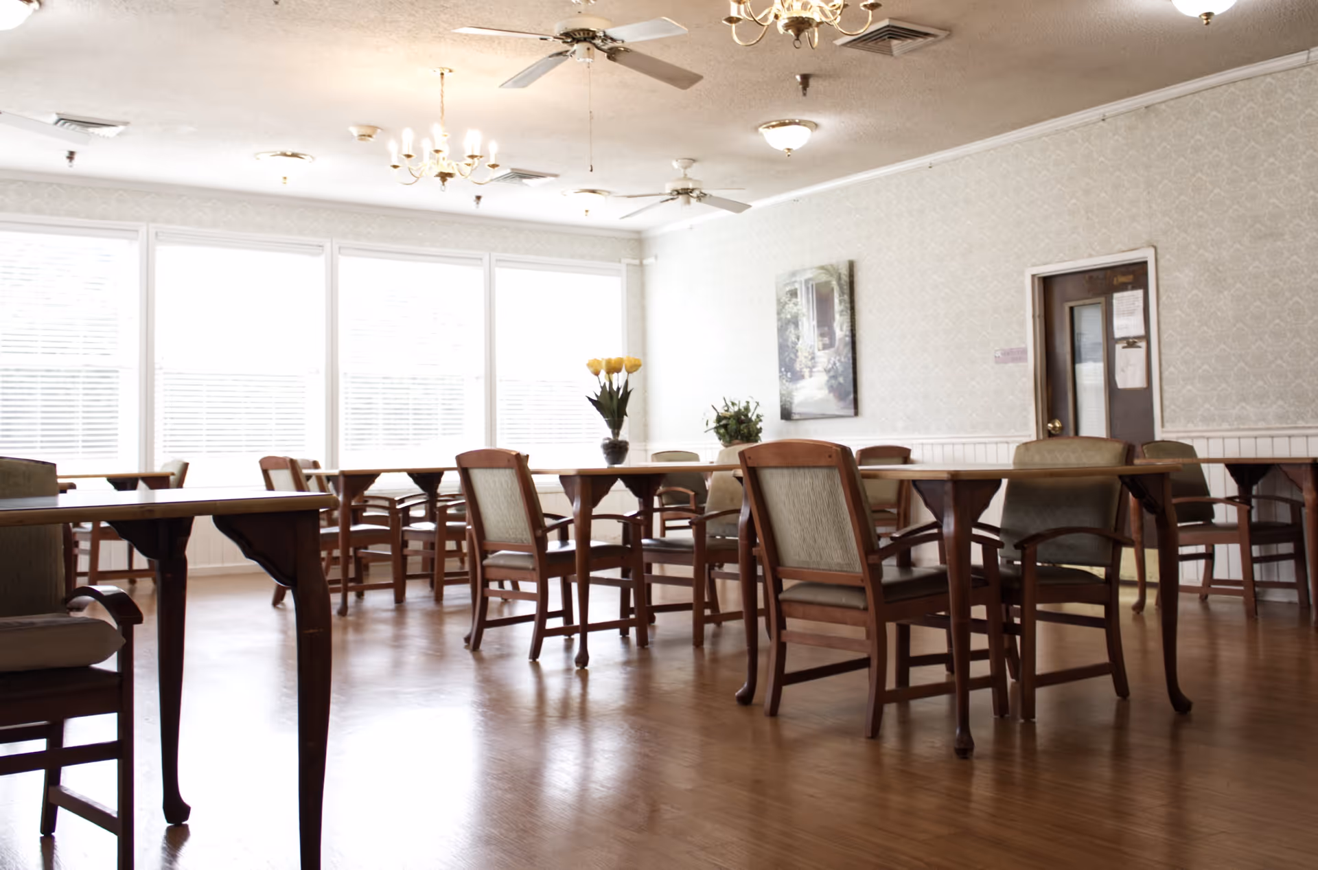 A bright dining room with wooden tables and chairs arranged neatly. The room has large windows with blinds allowing natural light to fill the space. There are ceiling fans and chandeliers on the ceiling, and a painting on the wall. Two vases with flowers are placed on the tables.