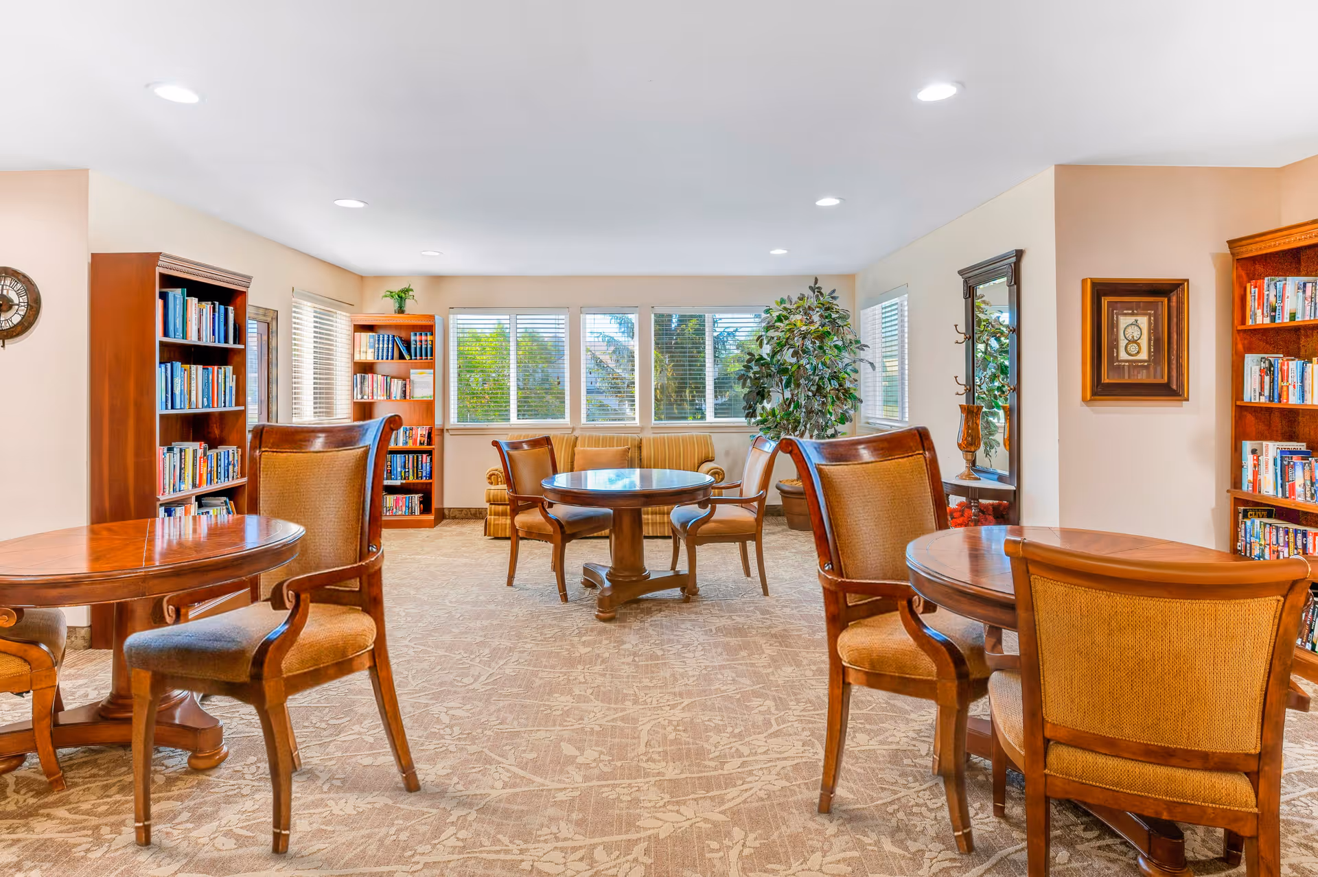 A bright and cozy common area with several wooden tables and cushioned chairs arranged on a patterned carpet. The room features large windows with white blinds letting in natural light, two wooden bookshelves filled with books, a small sofa, a large potted plant, a wall clock, and a mirror with a decorative vase beneath it.