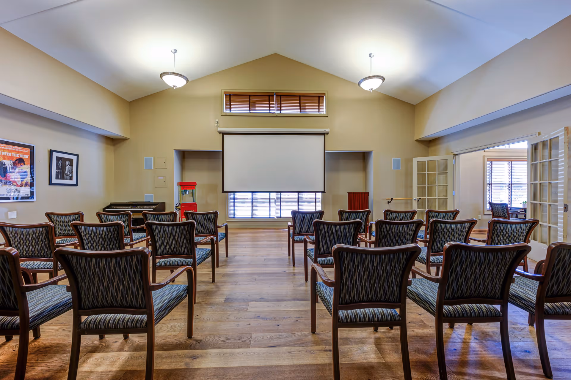 A small auditorium or meeting room with rows of wooden chairs facing a blank projector screen. The room has wooden flooring, beige walls, two ceiling lights, and a popcorn machine near the front. There are framed pictures on the left wall and glass double doors on the right side.