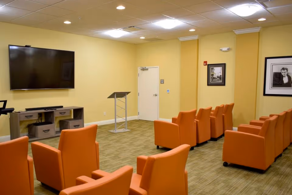 A small conference or presentation room with rows of orange armchairs facing a large flat-screen TV mounted on a yellow wall. There is a wooden media console below the TV and a metal podium near a closed white door. The room has framed black and white photographs on the walls and recessed ceiling lights.