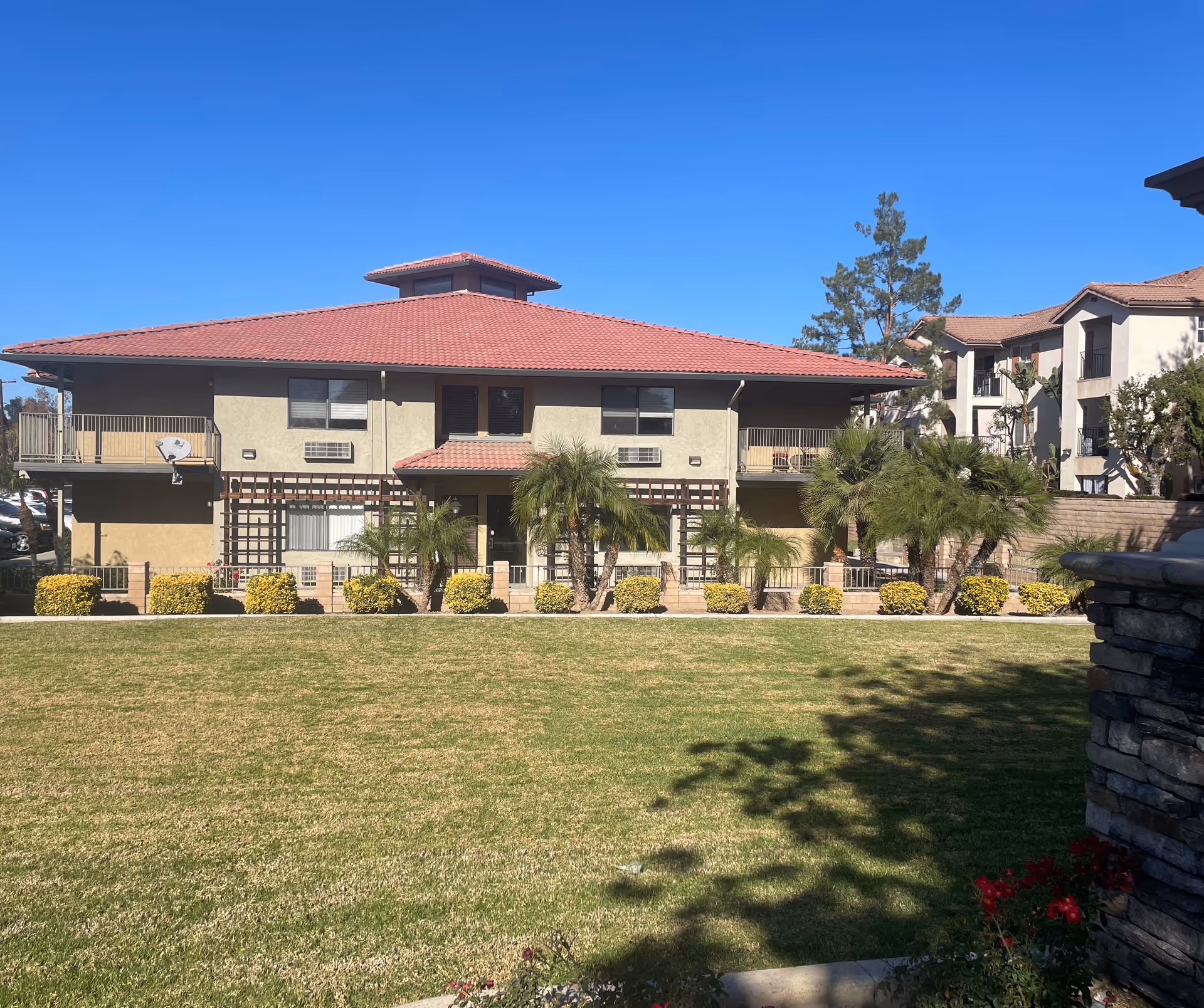 Exterior view of a two-story residential building with a red tile roof, beige walls, and balconies. The building is surrounded by a well-maintained lawn, palm trees, and shrubs under a clear blue sky.