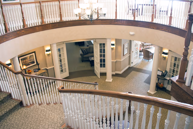 Interior view of a two-story lobby with a curved wooden railing and carpeted staircase overlooking seating areas and a chandelier.