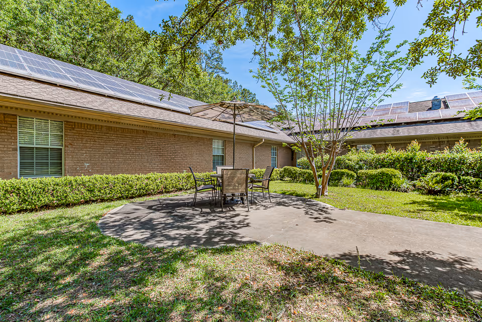 Patio area with a table, chairs and umbrella on a concrete pad beside a brick building with solar panels and surrounding greenery.