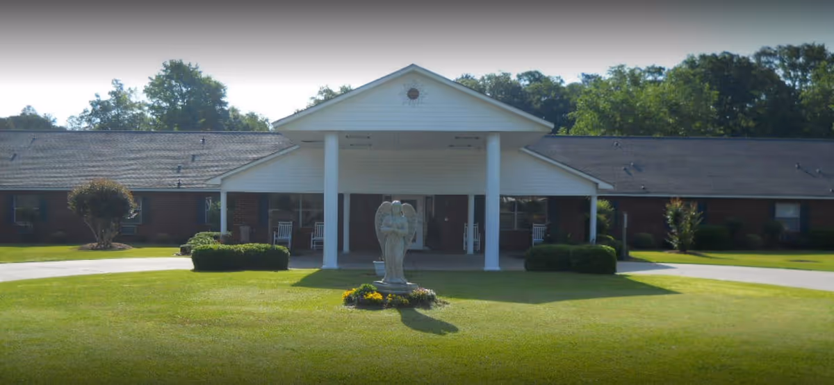 Front exterior view of a single-story brick building with a white covered entrance supported by two columns. In front of the entrance is a statue of an angel surrounded by a small flower bed. The building is surrounded by a well-maintained lawn and some bushes, with trees in the background.