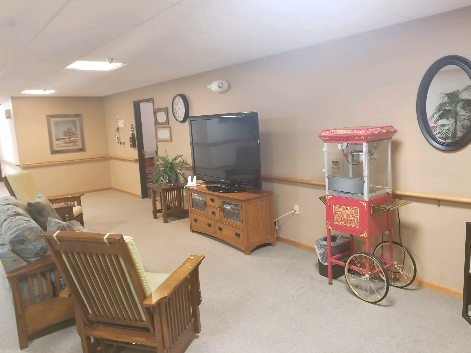 A cozy common area in a retirement community featuring wooden chairs with cushions, a wooden TV stand with a flat-screen television, a vintage-style red popcorn machine on wheels, a round wall mirror, a wall clock, a small side table with a plant, and a framed picture on the wall.