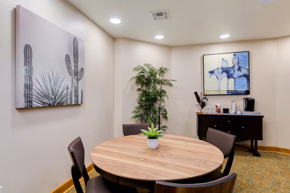 A small dining area with a round wooden table surrounded by four dark chairs. On the table is a small potted plant. The walls are light-colored and decorated with two framed artworks: one featuring cacti and another abstract painting in blue tones. In the corner, there is a tall green plant. Against the wall is a dark wooden sideboard with a coffee maker, glasses, and other small items on top. The room has recessed ceiling lights and patterned carpet flooring.