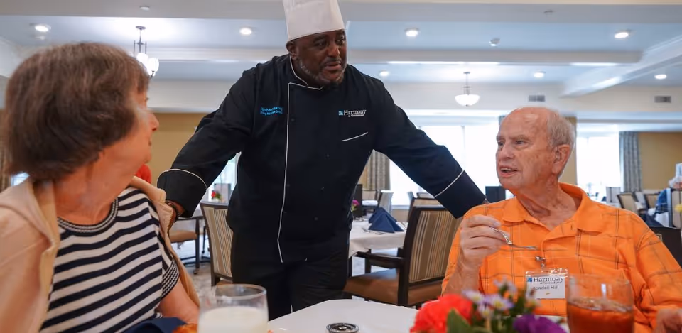 A chef speaks with two older residents seated at a table in a bright communal dining room.