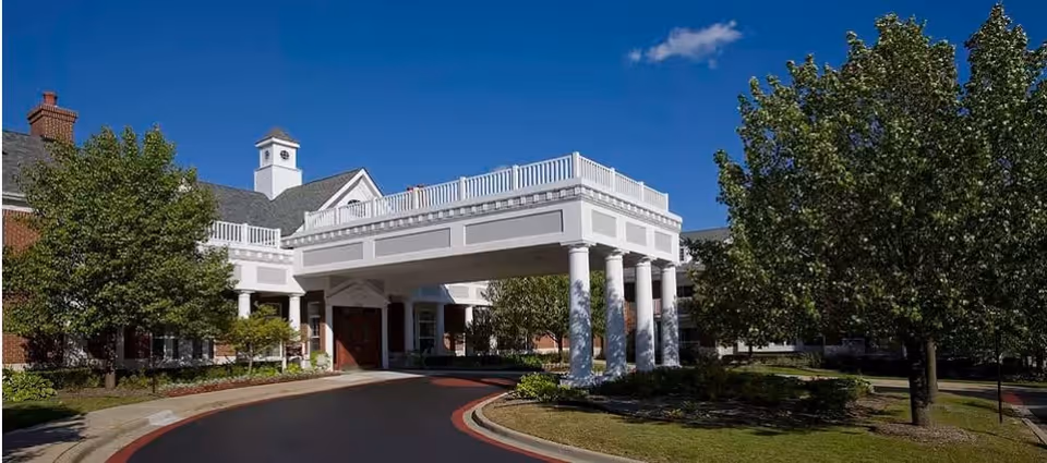 Exterior view of a senior living facility with a covered entrance supported by white columns, surrounded by trees and landscaping under a clear blue sky.