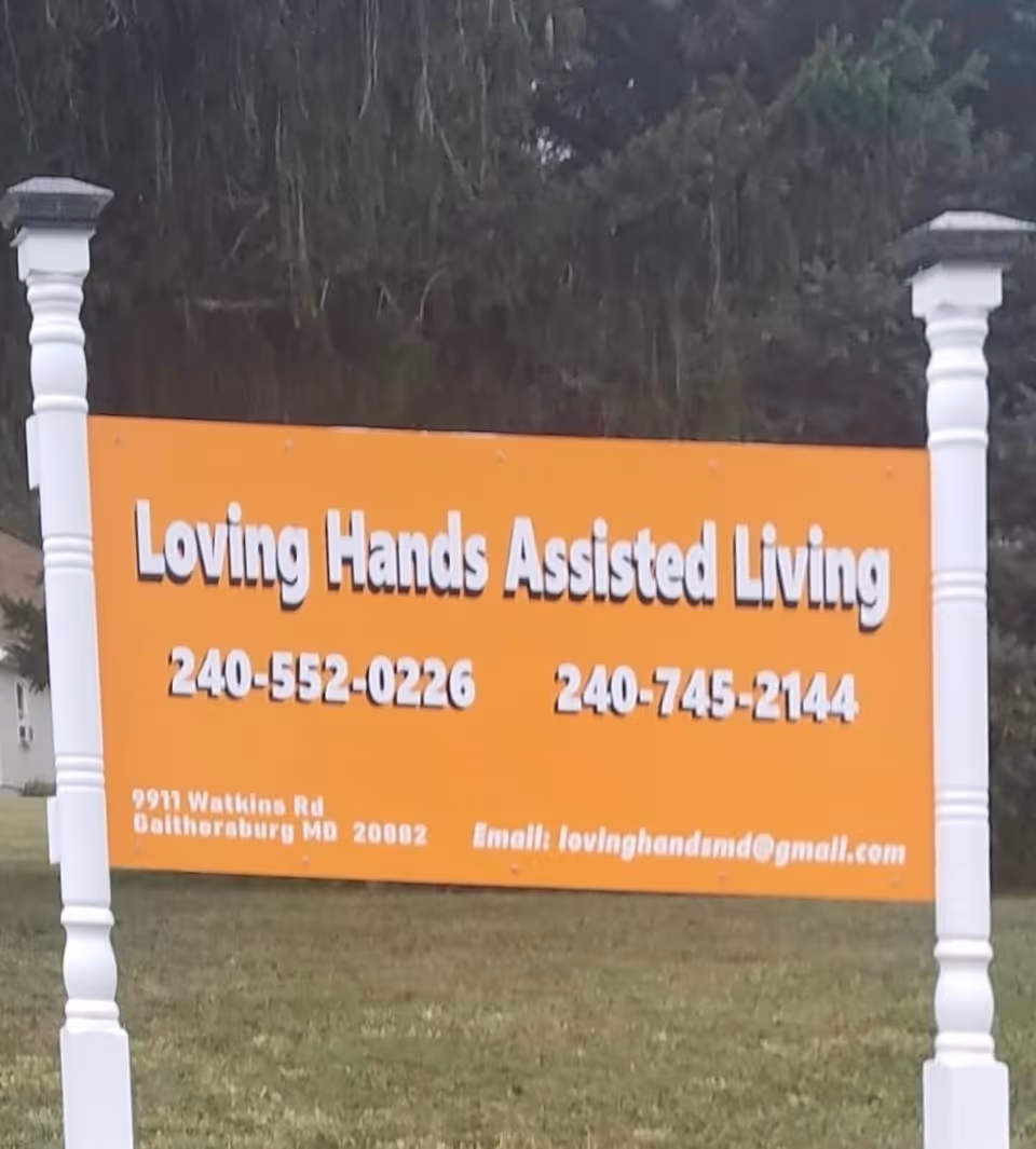 Orange lawn sign reading 'Loving Hands Assisted Living' with phone numbers and contact email mounted between two white posts.