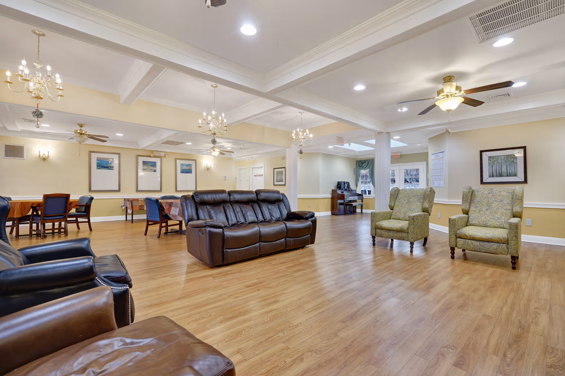A spacious and well-lit common area in a senior living facility featuring a mix of leather and upholstered armchairs and sofas arranged on a wood floor. The room has cream-colored walls adorned with framed artwork, chandeliers and ceiling fans providing lighting, and several tables with chairs in the background near the walls.