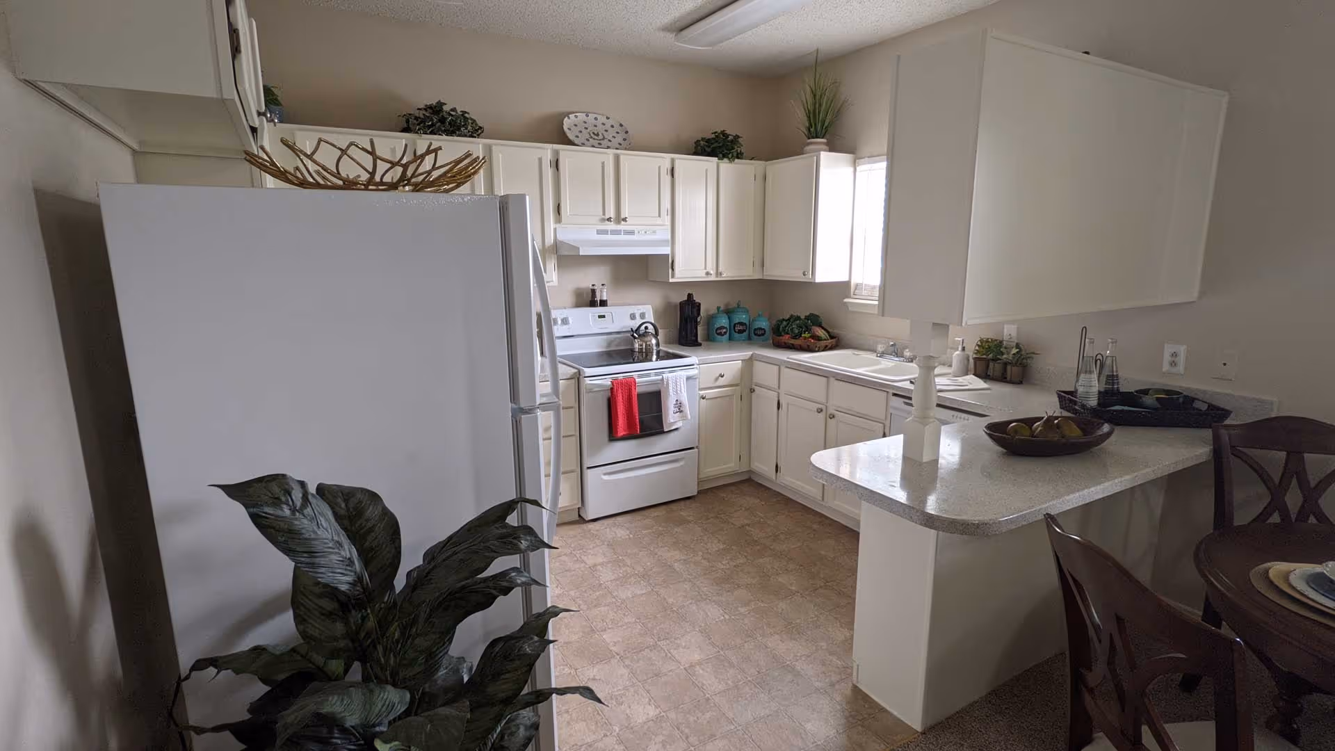A bright kitchen with white cabinets, a white refrigerator, stove, and dishwasher. The countertop has a bowl of fruit and decorative items. There is a small dining area with wooden chairs and a table partially visible on the right. A green leafy plant is in the foreground on the left side.