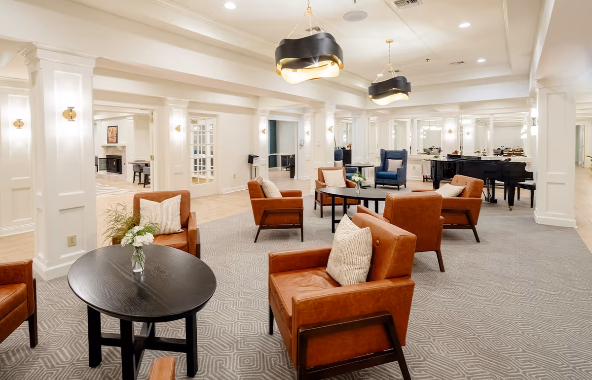 Bright, spacious senior living common lounge with leather armchairs, round tables, chandeliers and a grand piano.