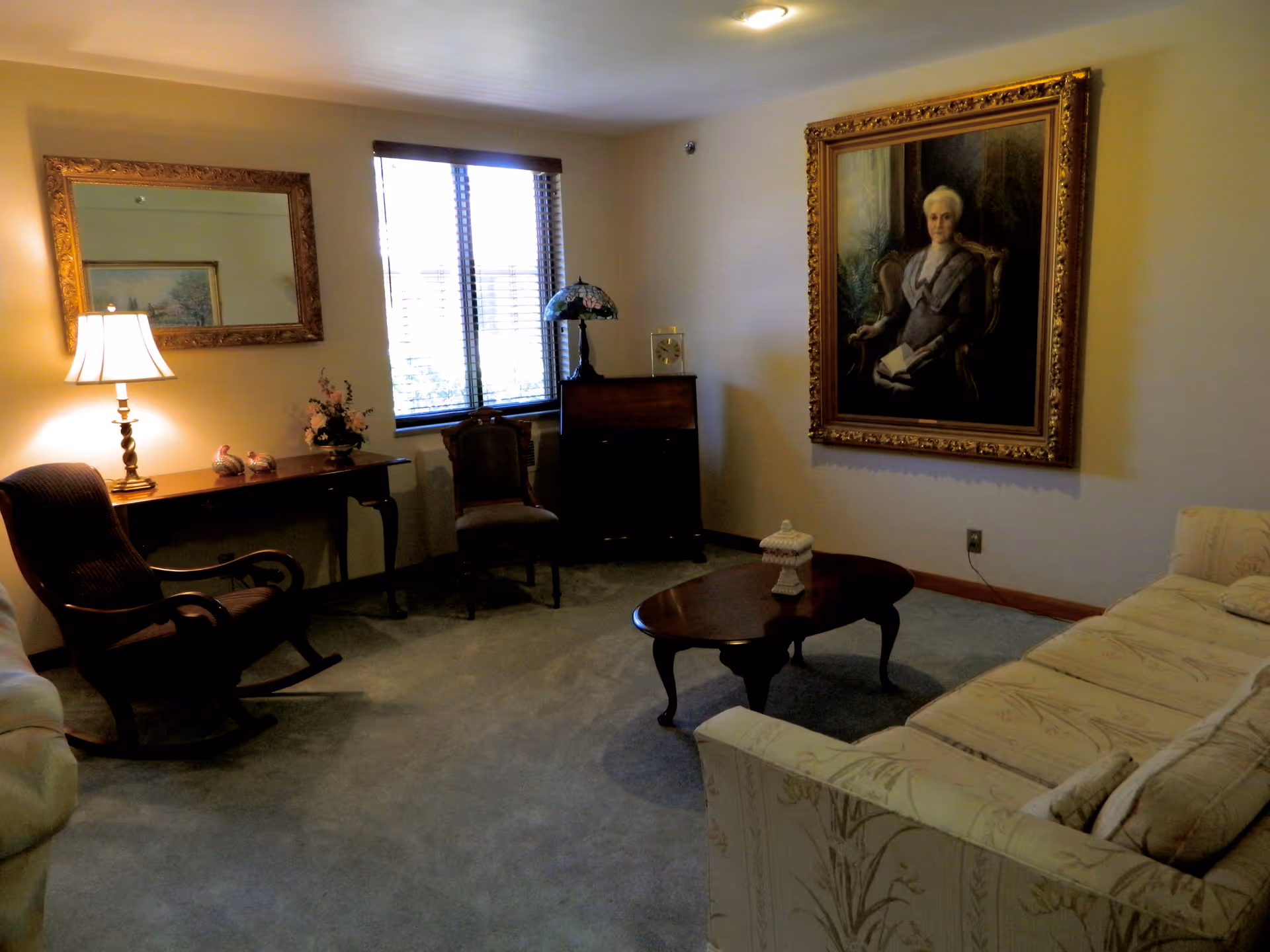 Cozy living room with an upholstered sofa, wooden coffee table and rocking chair, a side table with lamp, and a large framed portrait on the wall.