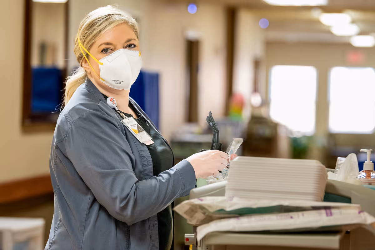 A healthcare worker wearing a face mask and gloves is standing next to a medical cart in a hallway of a healthcare facility, preparing medical supplies.