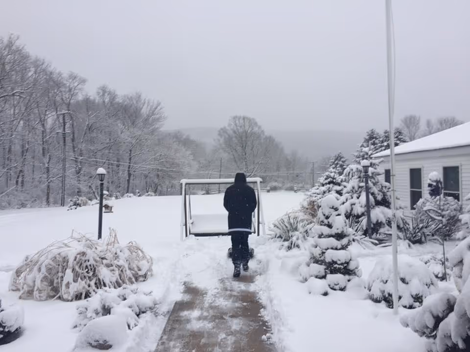 A person dressed in black winter clothing walking away on a snow-covered path in a snowy outdoor area with snow-covered bushes, trees, and a white building on the right side.