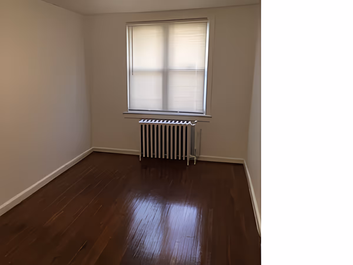 Empty room with polished wooden floor, beige walls, a window with a closed blind, and a radiator beneath the window.