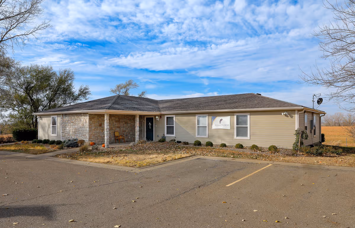 Single-story building with beige siding and a stone facade on the front porch area, surrounded by a parking lot and some trees under a partly cloudy blue sky. A sign on the building reads 'Kelly House'.