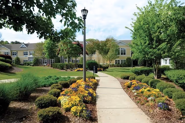 Landscaped walkway lined with flowers and shrubs leading to a two-story senior living building under a partly cloudy sky.